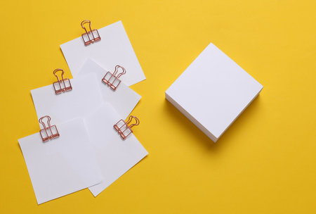 Memo white square sheets of paper with binder clips on yellow background. Stationery, education,...