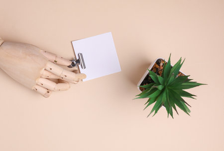 Wooden hand of puppet holding square memo card with metal binder on beige background