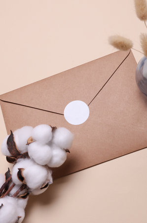 Kraft envelope with white blank sticker and cotton flowers on beige background.