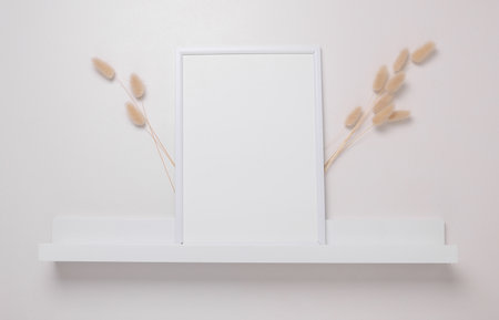Shelf with empty photo frame and dry flowers on white wall. Mockup for design