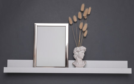 Shelf with white empty photo frame, bust vase with dry flowers on dark gray background. Mockup for design. Wall decorの写真素材