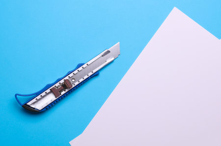 Metal stationery knife and white sheets of paper on a blue backgroundの写真素材