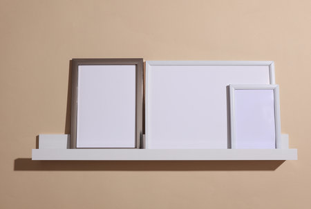 Shelf with white empty photo frames on beige background. Mockup for design. Wall decor