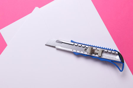 Metal stationery knife and white sheets of paper on a pink background