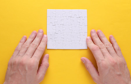Hands holding white blank assembled square jigsaw puzzle on yellow background. Mockup for designの写真素材