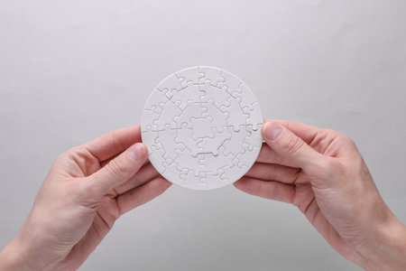 Hand holding white blank assembled round jigsaw puzzle on gray background. Mockup for design