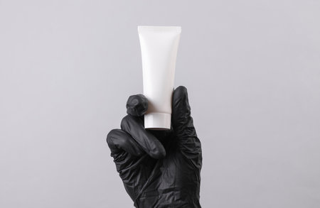 Hand of beautician in glove holds white empty tube of cream on gray background. Beauty concept. Mockup for designの写真素材