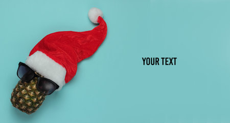 Creative New Year, Christmas concept. Pineapple dressed in glasses with Santa hat on blue background. Top view. Copy spaceの写真素材