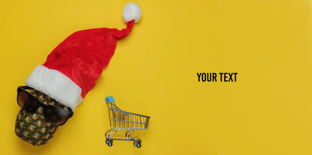 Christmas shopping. Pineapple dressed in Santa hat with sunglasses and shopping trolley on yellow background. Top view. Copy spaceの写真素材