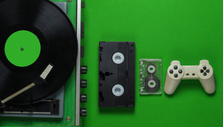 Flat lay retro media and entertainment. Vinyl record player with vinyl record, film camera, video cassette, audio cassette, gamepad on green background. 80s. Top viewの写真素材