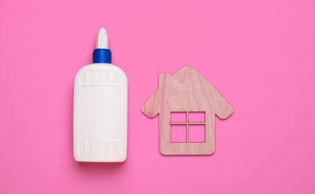 DIY house concept. Wooden mini house figurine and a bottle of glue on pink background. Top view