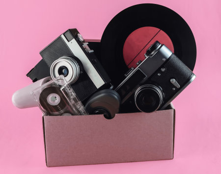 Cardboard box with retro film camera, gamepad, vinyl record, phone, audio cassette on pink background.の写真素材