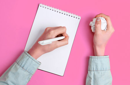 Lack of ideas. Female hands write in a notebook and hold a crumpled ball of paper on pink...