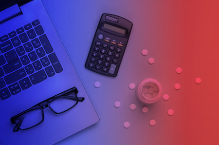 Economic calculation of the cost of treatment, analysis of the cost of medicines. Bottle of pills, laptop, calculator, glasses in neon light. Top viewの写真素材