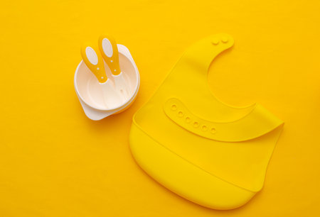 Silicone baby bib, bowl with spoon and fork on yellow background. Accessories for feeding a small childの写真素材