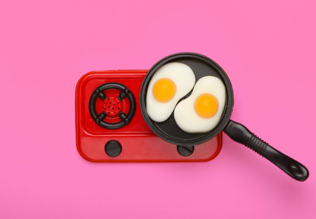Miniature toy frying pan with fried eggs on stove. Pink background. Top view. Minimalism. Studio shotの写真素材