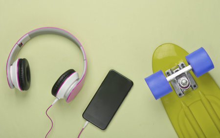 Flat lay hipster composition. Cruiser board and stereo headphones with smartphone on green pastel...