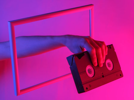 Creative art. Female hand holding video cassette through a soaring frame with neon holographic...