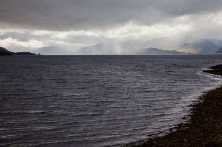 Scenic Scottish landscape with a silver grey lake and visible shore as well as a cloudy sky with sunny spells shining onto green mountains in backgroundの写真素材