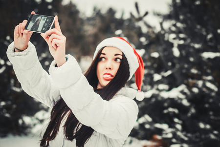 Girl taking selfie. Christmas girl outdoor self portrait. Woman in winter clothes and claus hat on a snow fieldの写真素材