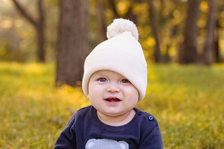 baby in white knitted hat sits on grass in Park against background of autumn trees