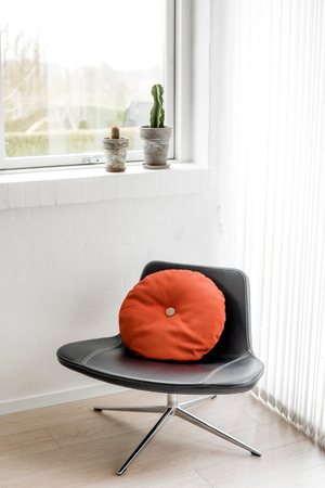 Black chair in a bright living room with a round orange pillow in retro design and cactus plants in the windowの写真素材