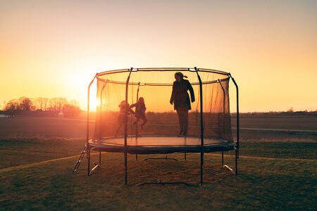 Trampoline on a lawn in the sunset with two kids and a young woman jumping and playingの写真素材