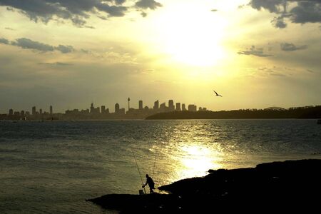 A silhouette fisheman tends to his poles at Watson's Bay with the Sydney skyline in the background.の写真素材