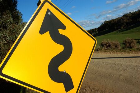 A windy road is warned by a big yellow sign showing a windy arrow.の写真素材