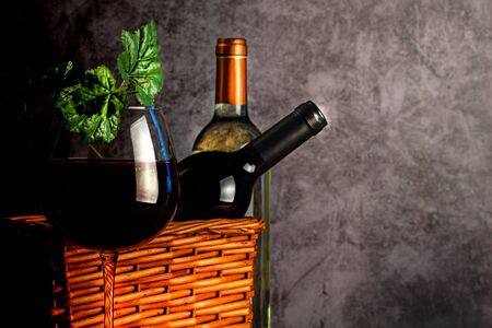 A bottle of wine in a basket and a glass on a thin leg. Grape leaves.の写真素材