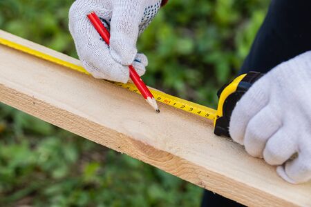 A carpenter in work gloves uses a pencil and construction tape measure to make marks on the Board....