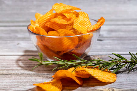 Potato chips with a sprig of rosemary in a glass bowl on a wooden table. A few slices are on the table.の写真素材