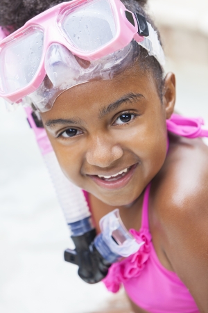 A cute happy young African American girl child relaxing on the side of a swimming pool smiling & wearing pink gogglesの写真素材