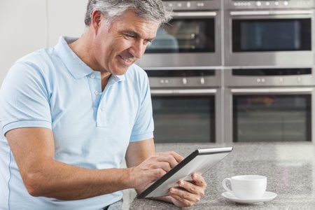 Middle aged man using tablet computer at home in the kitchen drinking coffee or teaの写真素材