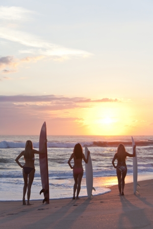 Rear view of three beautiful young woman surfer girls in bikinis with surfboard at a beach during sunset or sunriseの写真素材