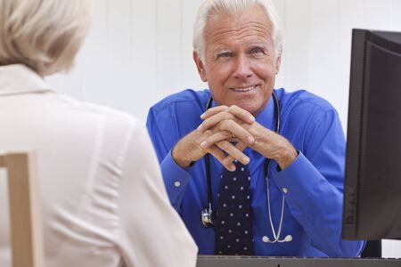 A smiling senior male medical doctor with a female patient and computer at his desk in an office at a hospitalの写真素材