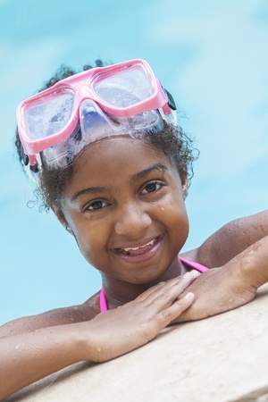 A cute happy young African American girl child relaxing on the side of a swimming pool wearing pink gogglesの写真素材
