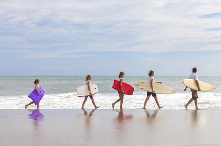 Rear view of family mother, father, daughter, parents and female girl children going surfing with...