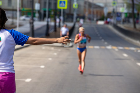 Hand volunteer girl with cup of water, in background female athlete running marathonの写真素材