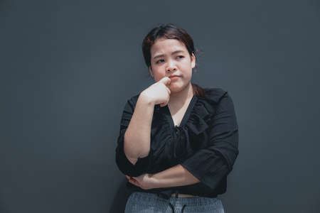 Young asian woman with chubby body put her finger on face looking away to think about something. Isolated on dark background with copy spaceの写真素材