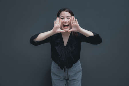 Excited young asian woman shouting with hands cupped around mouth isolated on grey background with copy spaceの写真素材