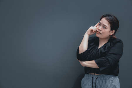 Young asian woman with chubby body put her finger on face looking away to think about something. Isolated on dark background with copy spaceの写真素材