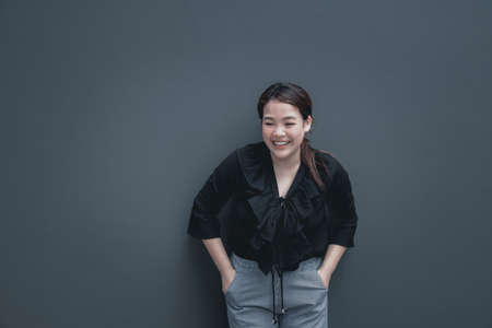 Young friendly face asian woman with chubby body have a happy smile put her hand on another arm. Back shirt with grey pants. Grey background with copy space.の写真素材