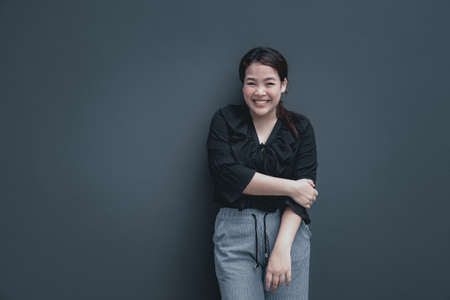 Young friendly face asian woman with chubby body have a happy smile put her hand on another arm. Back shirt with grey pants. Grey background with copy space.の写真素材