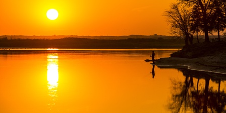 Golden orange sunset over the calm waters of the river.  The silhouette of a fisherman can be seen...