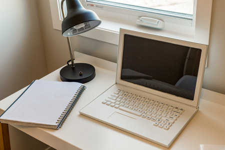 A laptop is open on a desk next to a notebook. The laptop is black and white, and the notebook is white. The desk is clean and organizedの写真素材