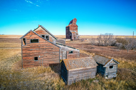 A small house with a barn in the background. The barn is old and the house is also old. The sky is blue and the grass is dryの写真素材