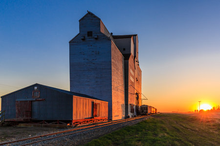 A train is pulling a large grain silo. The sun is setting in the background. The silo is old and the train is movingの写真素材