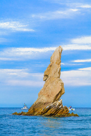 A large rock sits on the ocean shore. A boat is in the water near the rock. The sky is blue and cloudyの写真素材