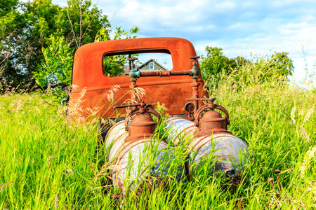 An old rusty truck is parked in a field of grass. The truck is surrounded by two tanks, one of which...